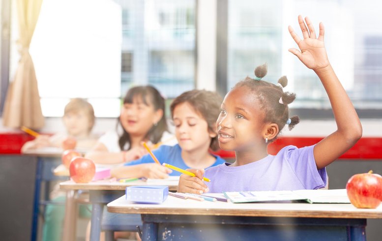 Des enfants dans une salle de classe, assis à des bureaux et levant la main pour répondre à une question. L'atmosphère est joyeuse et engageante, avec des pommes sur les bureaux et un éclairage naturel filtrant à travers les fenêtres.