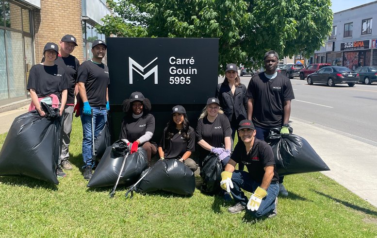 Groupe de bénévoles MACH souriants posant devant l’immeuble Carré Gouin, après une activité de nettoyage communautaire. Chacun tient des sacs de déchets remplis, illustrant leur engagement pour un environnement plus propre. Ils sont vêtus de t-shirts noirs portant le logo de l'entreprise, montrant leur enthousiasme et leur engagement dans cette initiative communautaire.