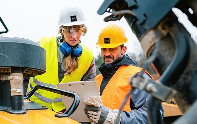 Deux travailleurs de construction de MACH en tenue de sécurité, portant des casques et des gilets de haute visibilité, discutent et examinent un document sur un chantier. L'un des travailleurs tient une tablette tandis que l'autre observe les informations.