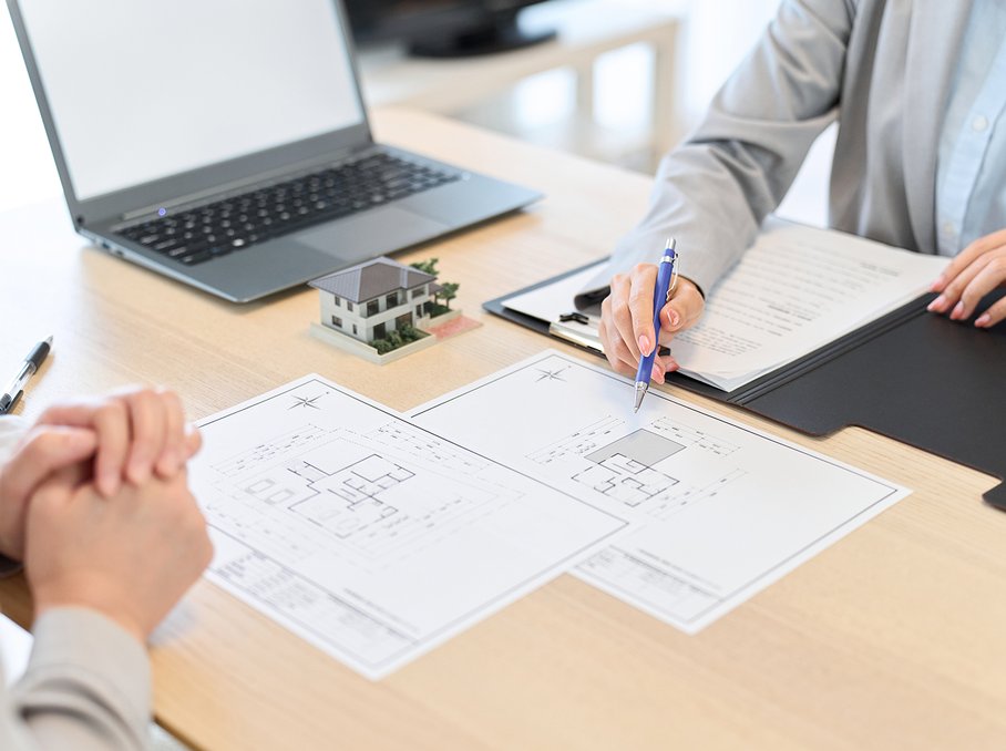 Two people sit at a table reviewing architectural plans, with a laptop and a small house model. The image represents a real estate brokerage or leasing meeting.