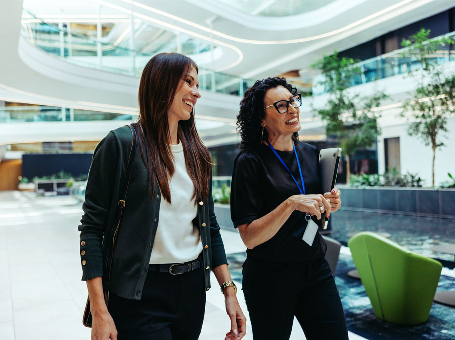 Two businesswomen walk side by side through a bright, modern building atrium, smiling and engaged in conversation. The setting is contemporary, featuring greenery, large windows, and green seating.