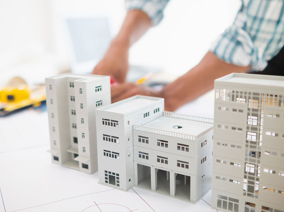 Maquettes d’immeubles modernes posées sur des plans d’architecture, avec une personne en arrière-plan travaillant sur un bureau. L’image illustre la conception et le développement immobilier.