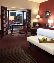 Elegant hotel room with a large double bed, décor in burgundy and cream tones, a cozy sitting area, and a large window overlooking downtown skyscrapers.